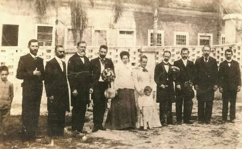 Fotografia em preto e branco do início do século XX, mostrando um grupo de pessoas — homens, mulheres e uma criança — posando em frente ao prédio do Grupo Escolar Dr. Bernardino de Campos, em São Roque. Os adultos usam trajes formais da época, com vestidos longos e ternos escuros. Ao fundo, o edifício com janelas retangulares e elementos arquitetônicos característicos do período.