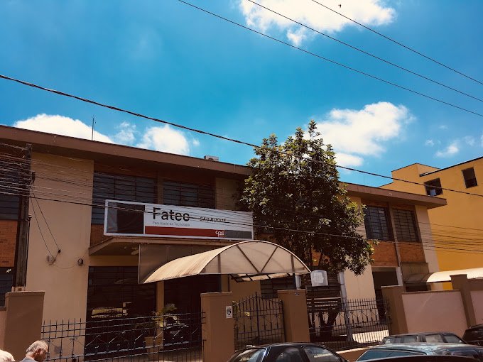 Fachada da FATEC São Roque vista em um dia ensolarado, com céu azul e poucas nuvens. A entrada principal possui uma marquise metálica curva sobre o portão e uma placa com a identificação “Fatec São Roque - Faculdade de Tecnologia” ao centro do prédio, que tem janelas amplas e um muro com gradil.
